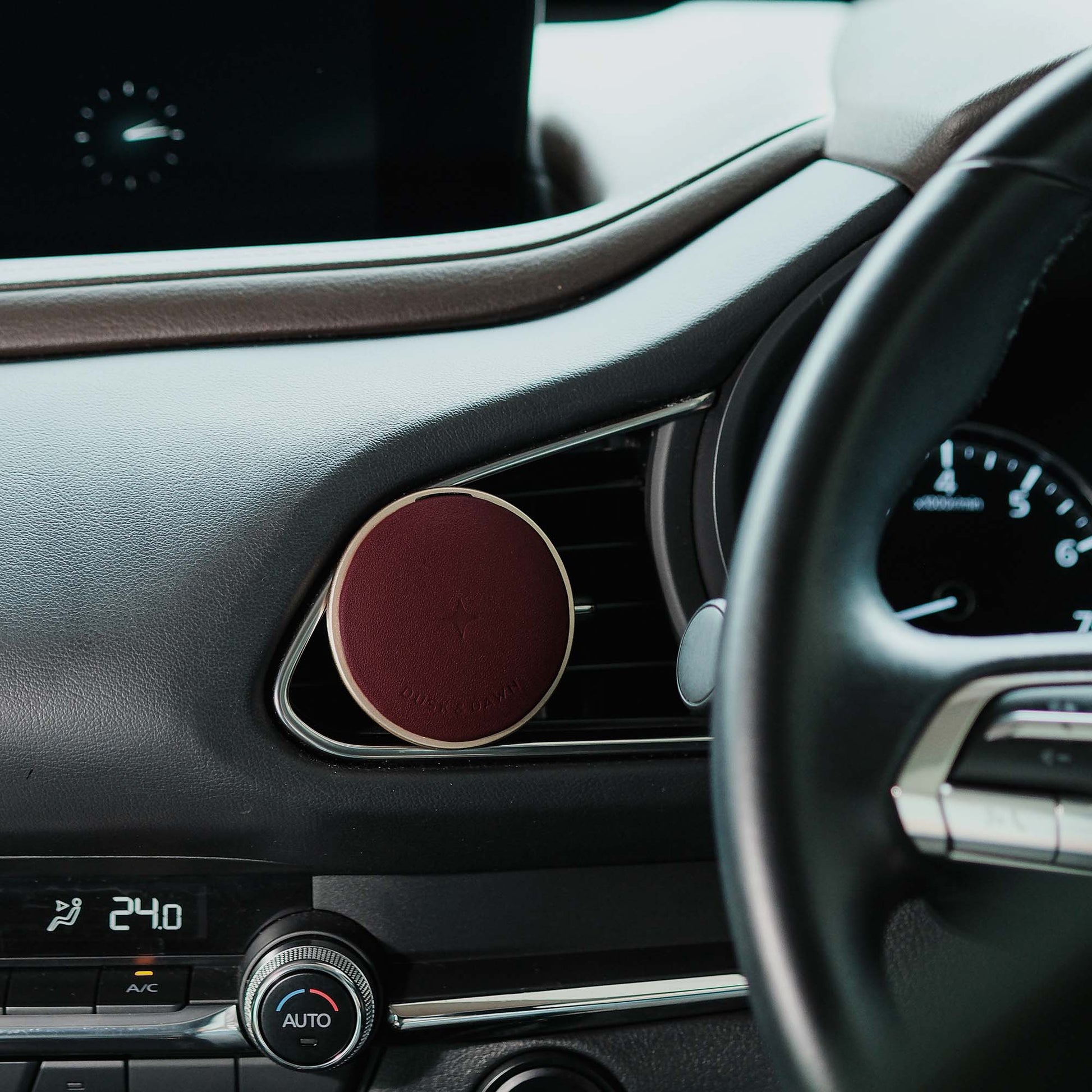 A round car air freshener diffuser with a deep burgundy red center and a light beige outer rim, mounted on a car's air vent dashboard.