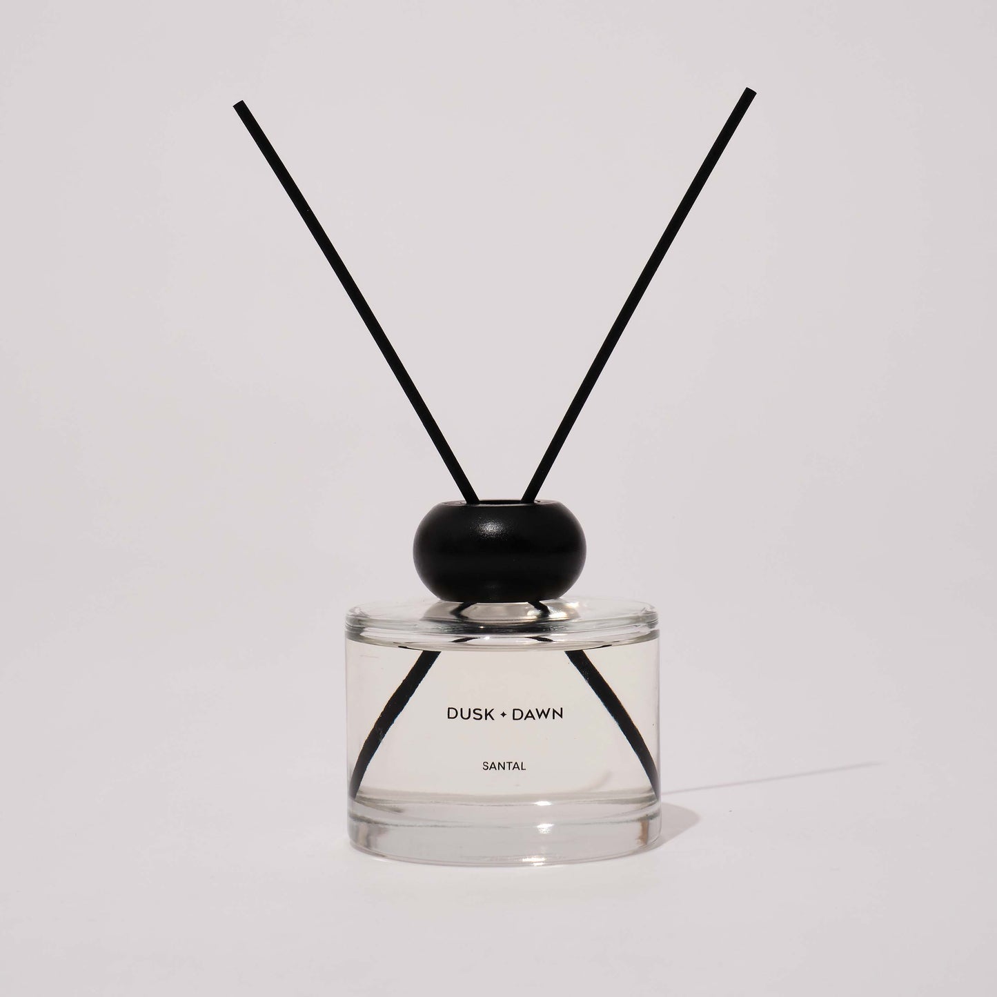 A reed diffuser bottle with clear glass, a black wooden cap, and two black reeds, labeled 'DUSK + DAWN' and 'SANTAL'.