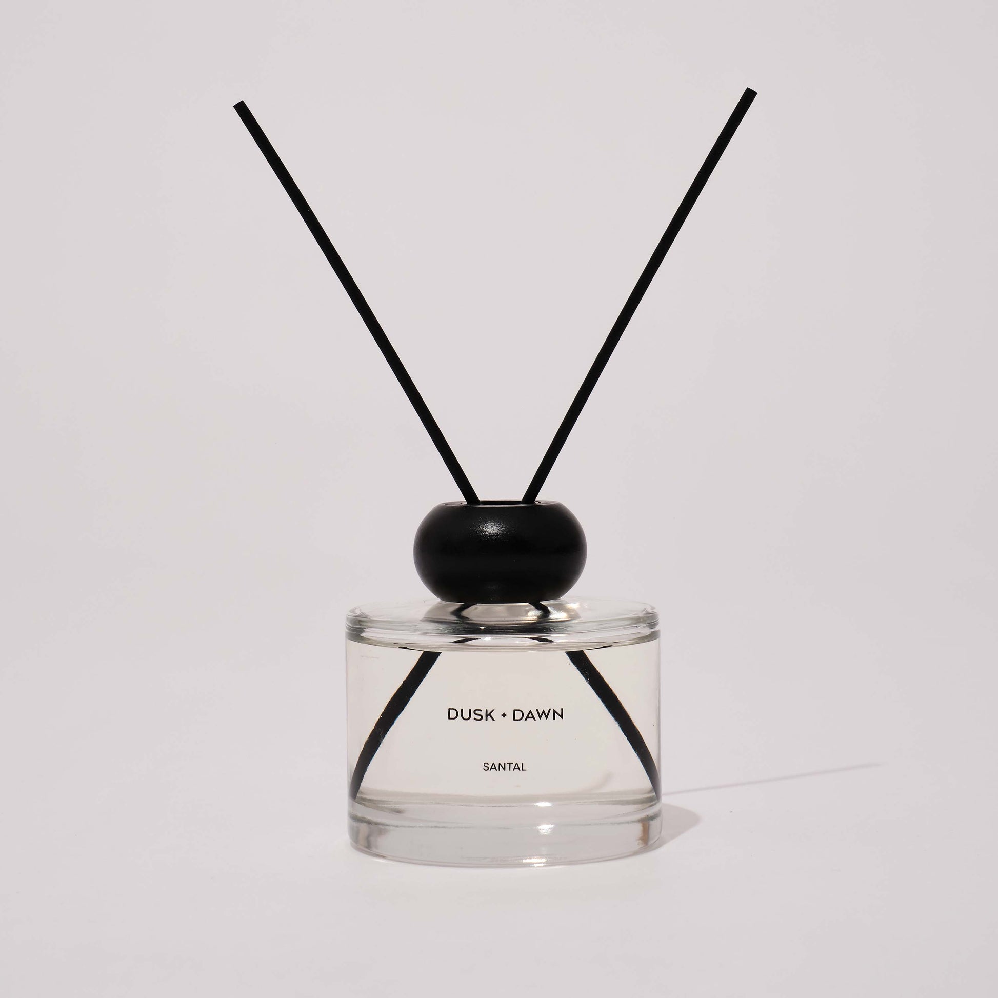 A reed diffuser bottle with clear glass, a black wooden cap, and two black reeds, labeled 'DUSK + DAWN' and 'SANTAL'.