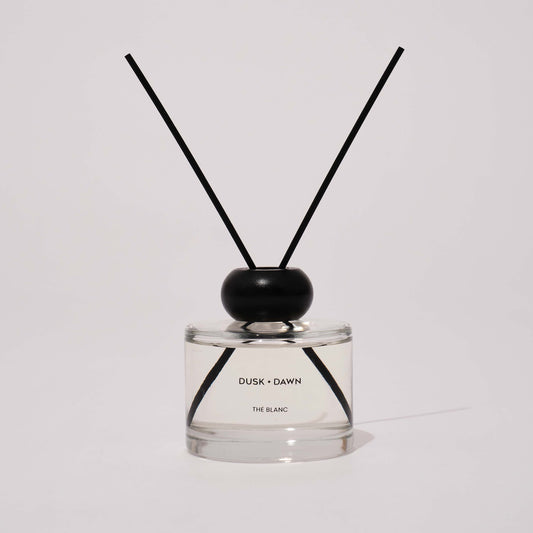 A reed diffuser bottle with clear glass, a black wooden cap, and two black reeds, labeled 'DUSK + DAWN' and 'THE BLANC'.