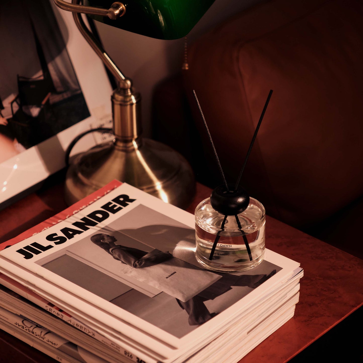 A clear glass reed diffuser bottle with black reeds sits on top of a stack of magazines on a wooden table, next to a brass desk lamp in a dimly lit room.